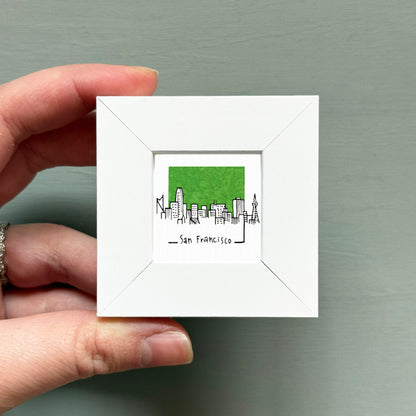 A hand is holding a small white square frame containing a green illustration of a city skyline.