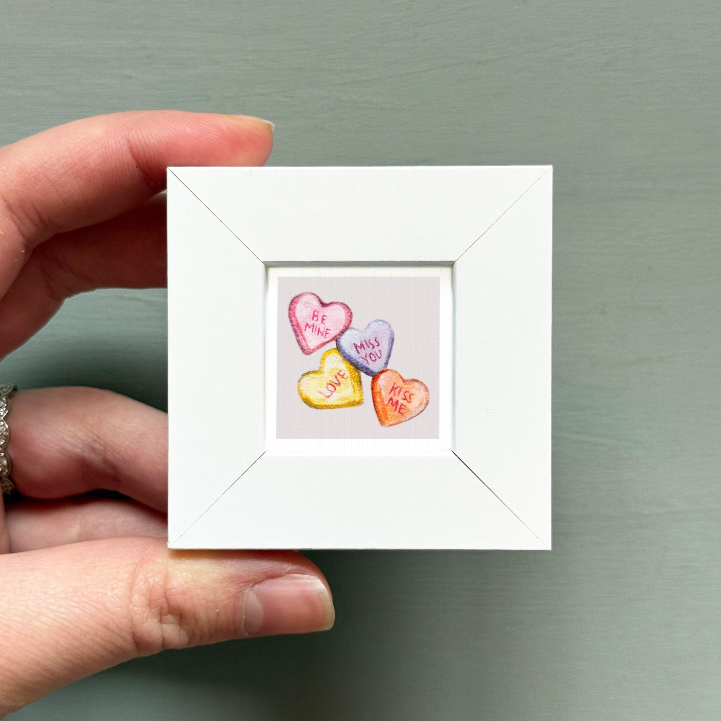 A hand is holding a small white picture frame containing a colorful heart-shaped candy with the words "Be Mine" written on it.
