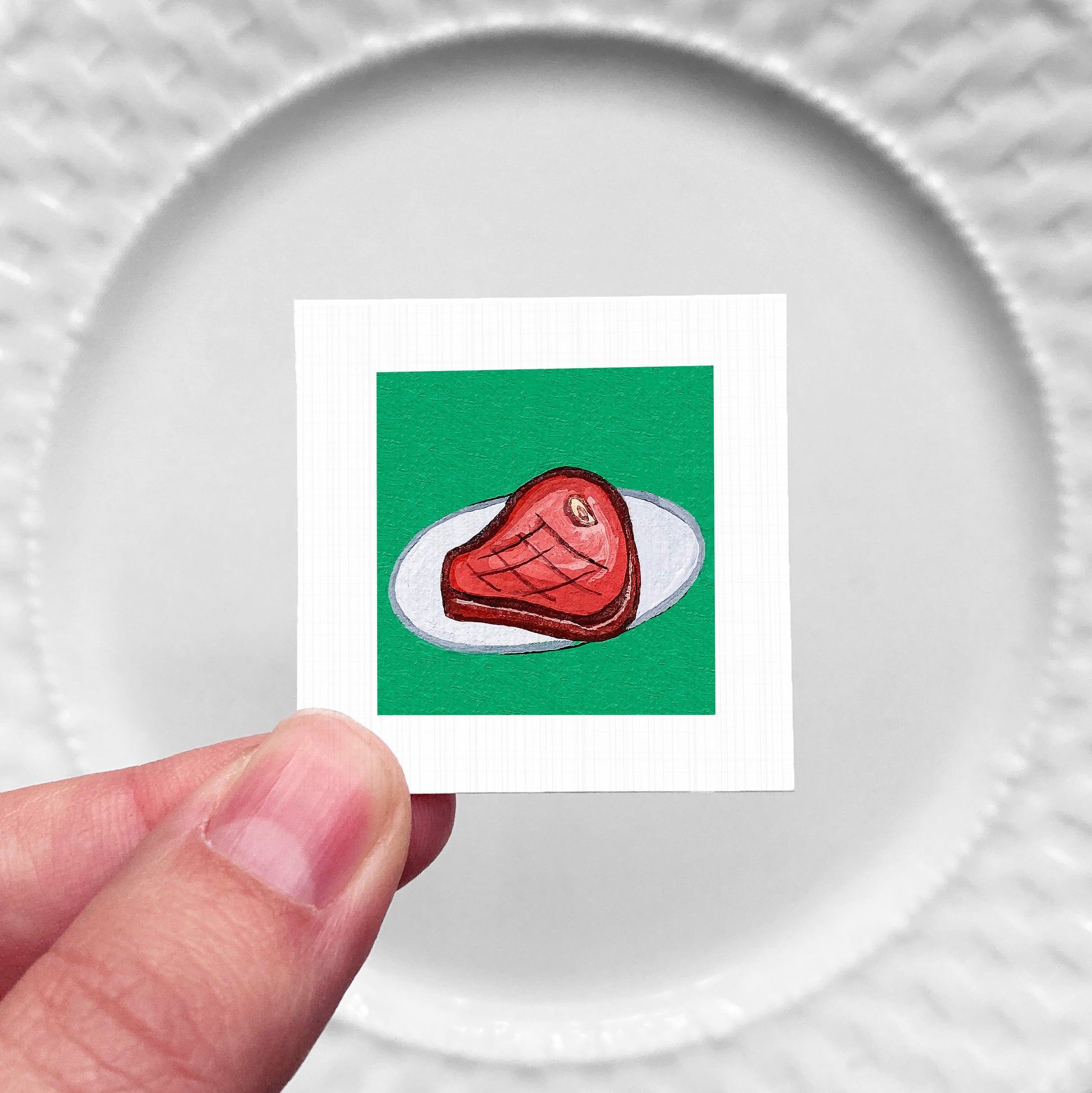 A hand is holding a small square with a picture of a piece of meat on a green background.