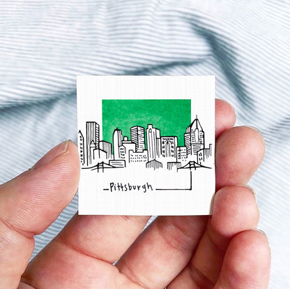 A hand holding a small square with a drawing of a city skyline and the word "Pittsburgh" written below it.