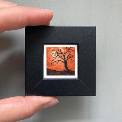A person's hand is holding a small, square-shaped black frame containing a miniature painting of a tree with an orange background and a full moon.