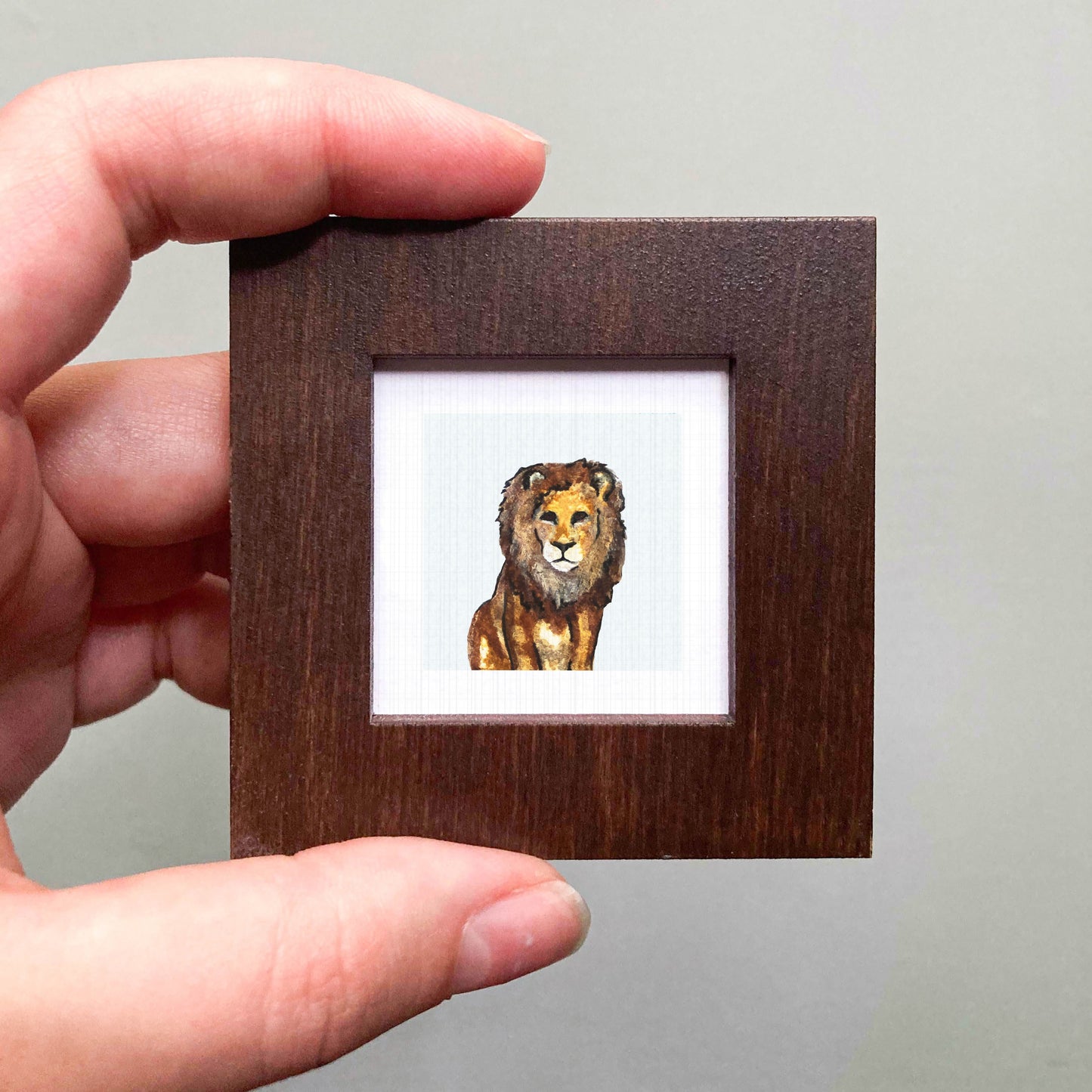 A hand is holding a small wooden frame containing a watercolor painting of a lion.