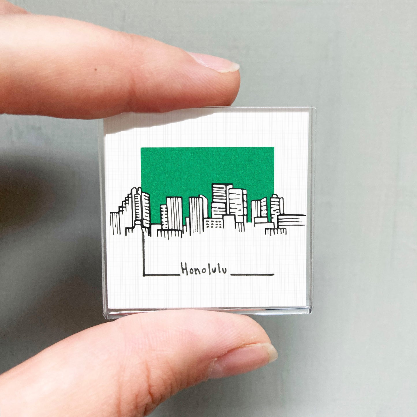 A person's hand holding a small square magnet with a green cityscape illustration on it.