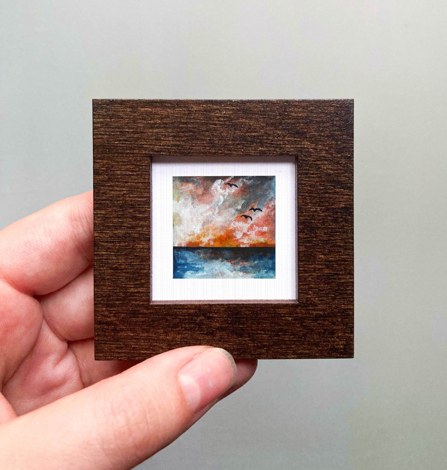 A hand is holding a small, square-shaped wooden frame containing a painting of a sunset over the ocean.