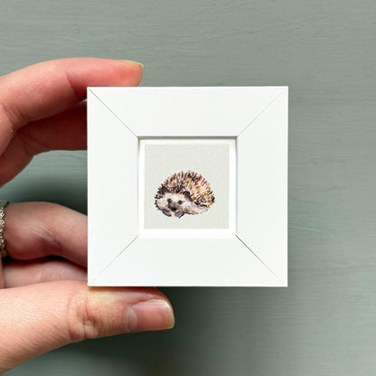 A hand is holding a small white picture frame containing a painting of a hedgehog.