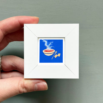 A hand is holding a small white square frame containing an illustration of a bowl of soup with steam rising from it.