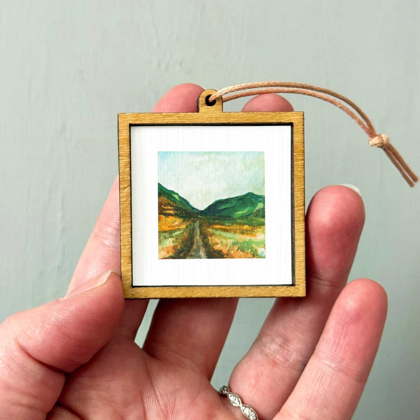 A hand holding a small wooden frame containing a painting of a landscape with mountains and a winding road.