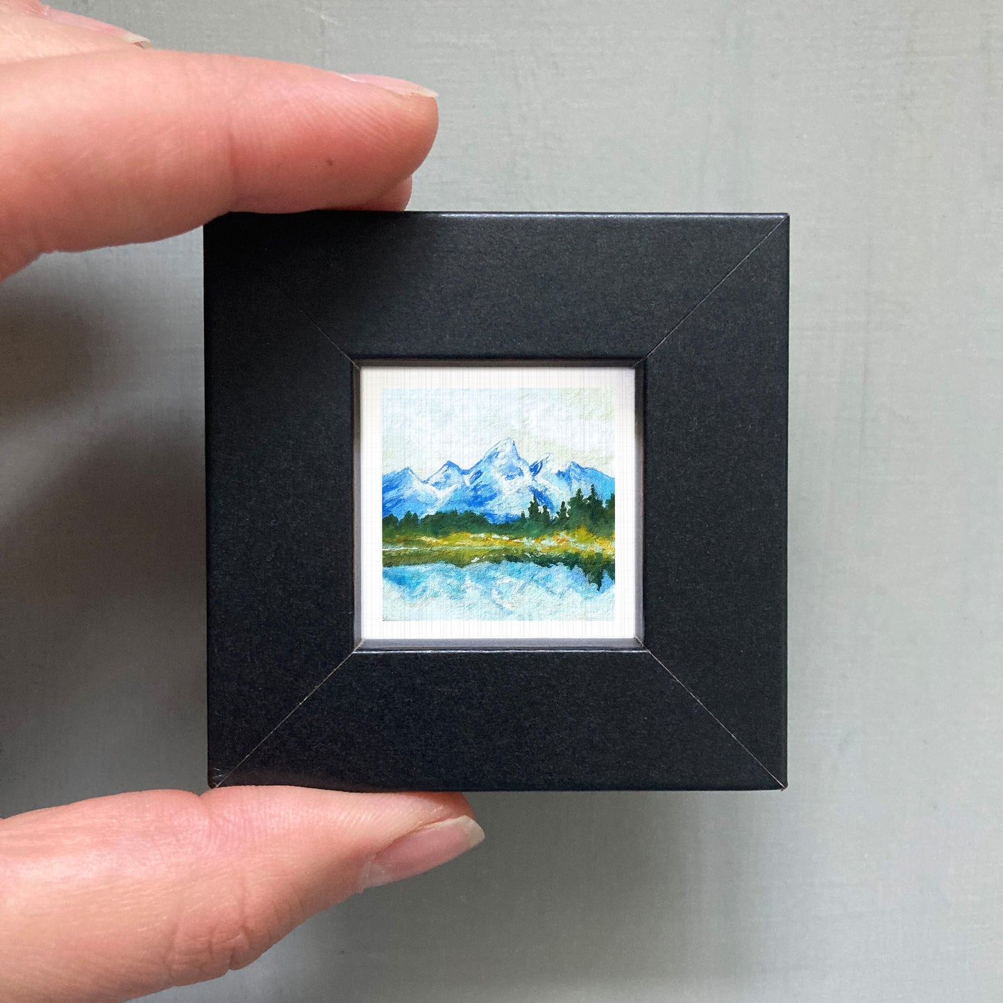 A person's hand holding a small black picture frame containing a landscape painting of a mountain scene.