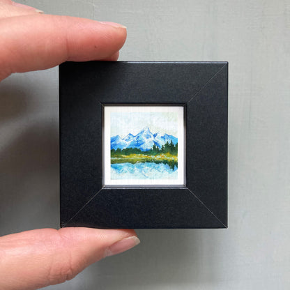 A person's hand holding a small black picture frame containing a landscape painting of a mountain scene.