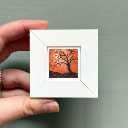 A hand is holding a small white square frame containing a painting of a tree with a full moon in the background.
