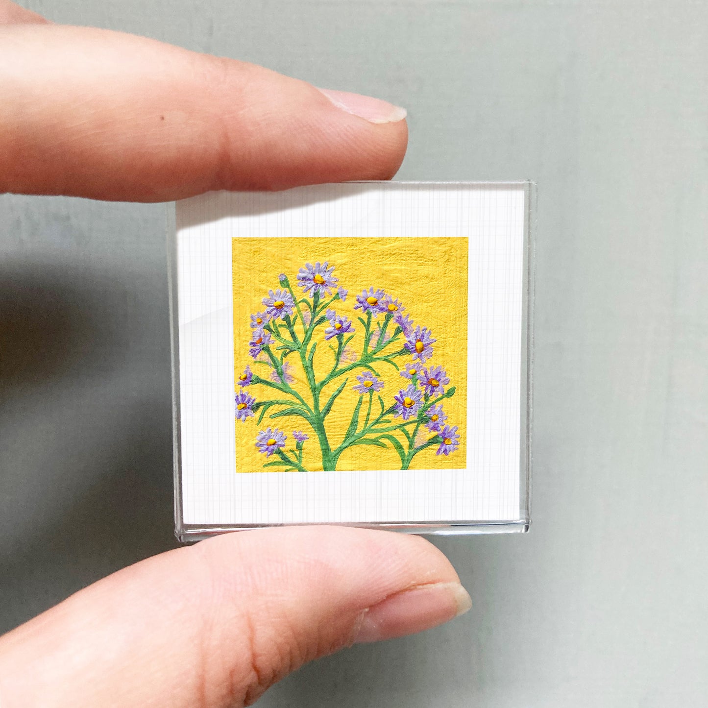 A person's hand is holding a small square picture of a yellow flower with purple petals.