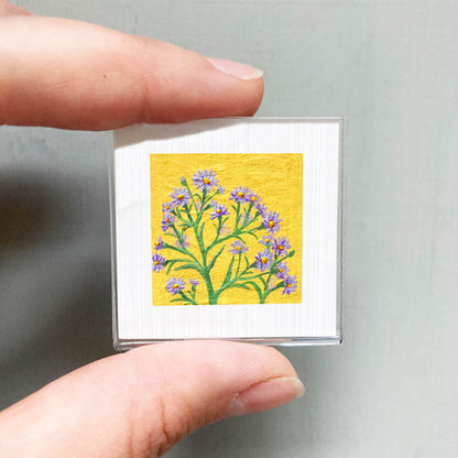 A person's hand is holding a small square picture of a yellow flower with purple petals.