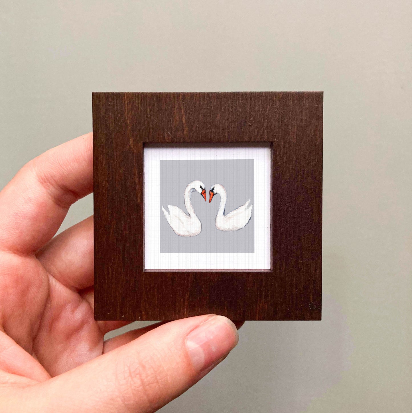 A hand is holding a small wooden frame containing a picture of two white swans with their necks intertwined.