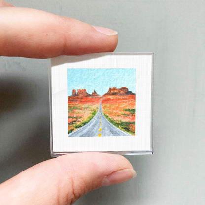 A person's hand holding a small square picture depicting a desert landscape with a winding road leading to distant mountains.