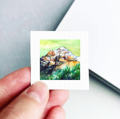 A hand holding a small square artwork depicting a mountain landscape with green and brown tones.