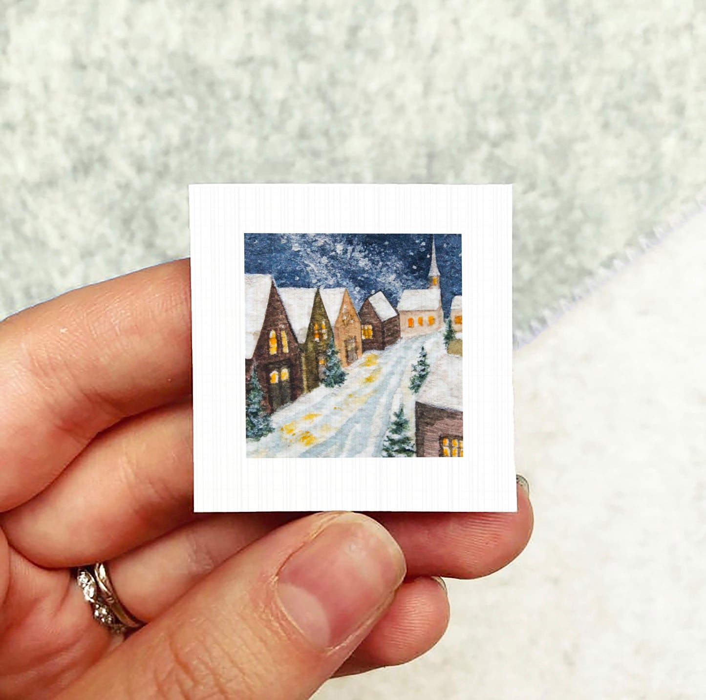 A hand holding a small square picture depicting a snowy village scene with houses and trees.