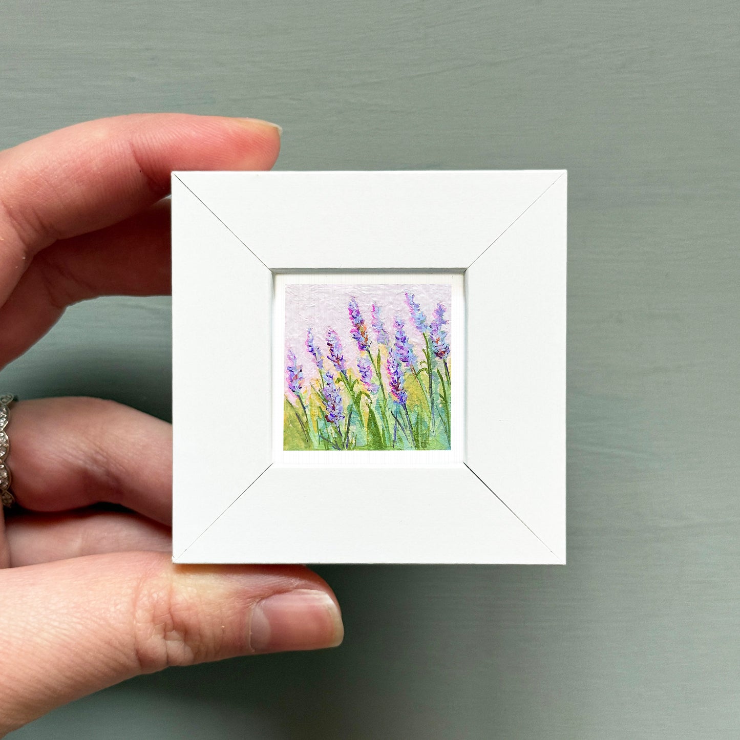 A hand is holding a small white square frame containing a painting of purple flowers.