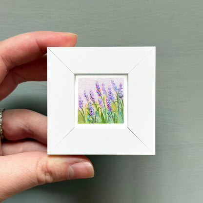 A hand is holding a small white square frame containing a painting of purple flowers.