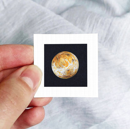 A hand is holding a small square photograph of a planet against a black background.