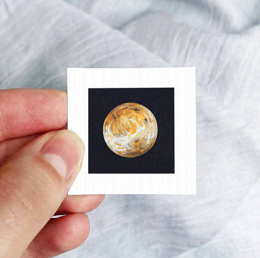 A hand is holding a small square photograph of a planet against a black background.