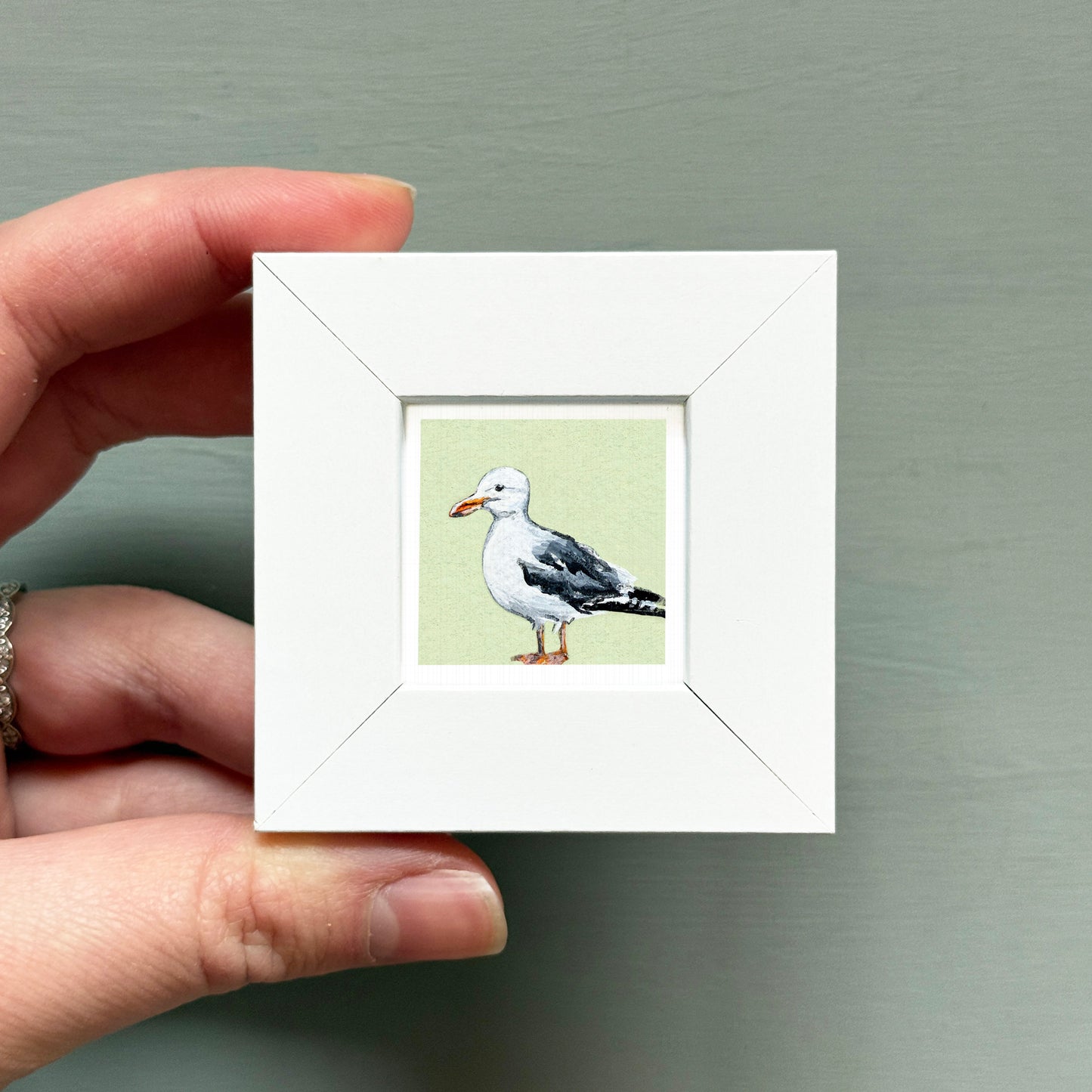A hand is holding a small white picture frame containing a painting of a seagull.