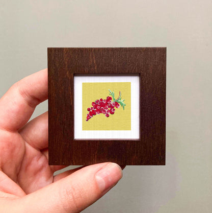 A hand is holding a small wooden frame containing a framed artwork of a bunch of red grapes.