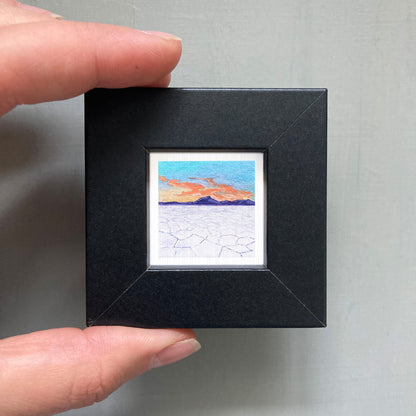 A person's hand is holding a small black picture frame containing a miniature painting of a desert landscape with a sunset in the background.
