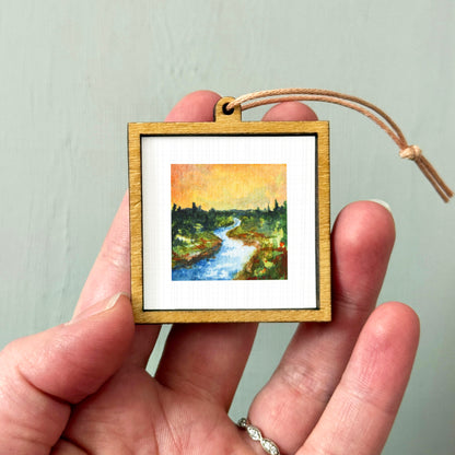 A hand holding a small wooden frame containing a painting of a river and trees.