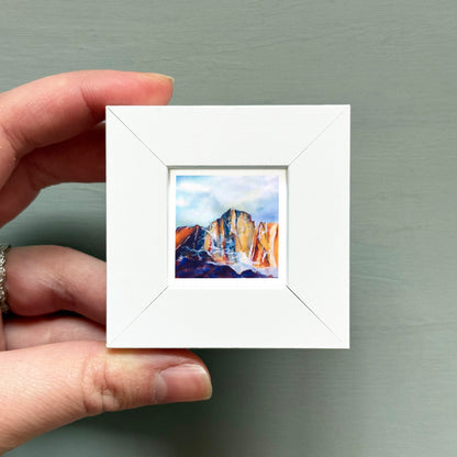 A hand is holding a small white square frame containing a watercolor painting of a mountain landscape.