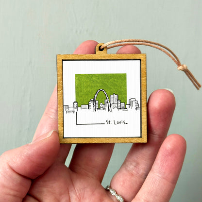 A hand holding a small wooden frame with a picture of St. Louis, Missouri, inside it.