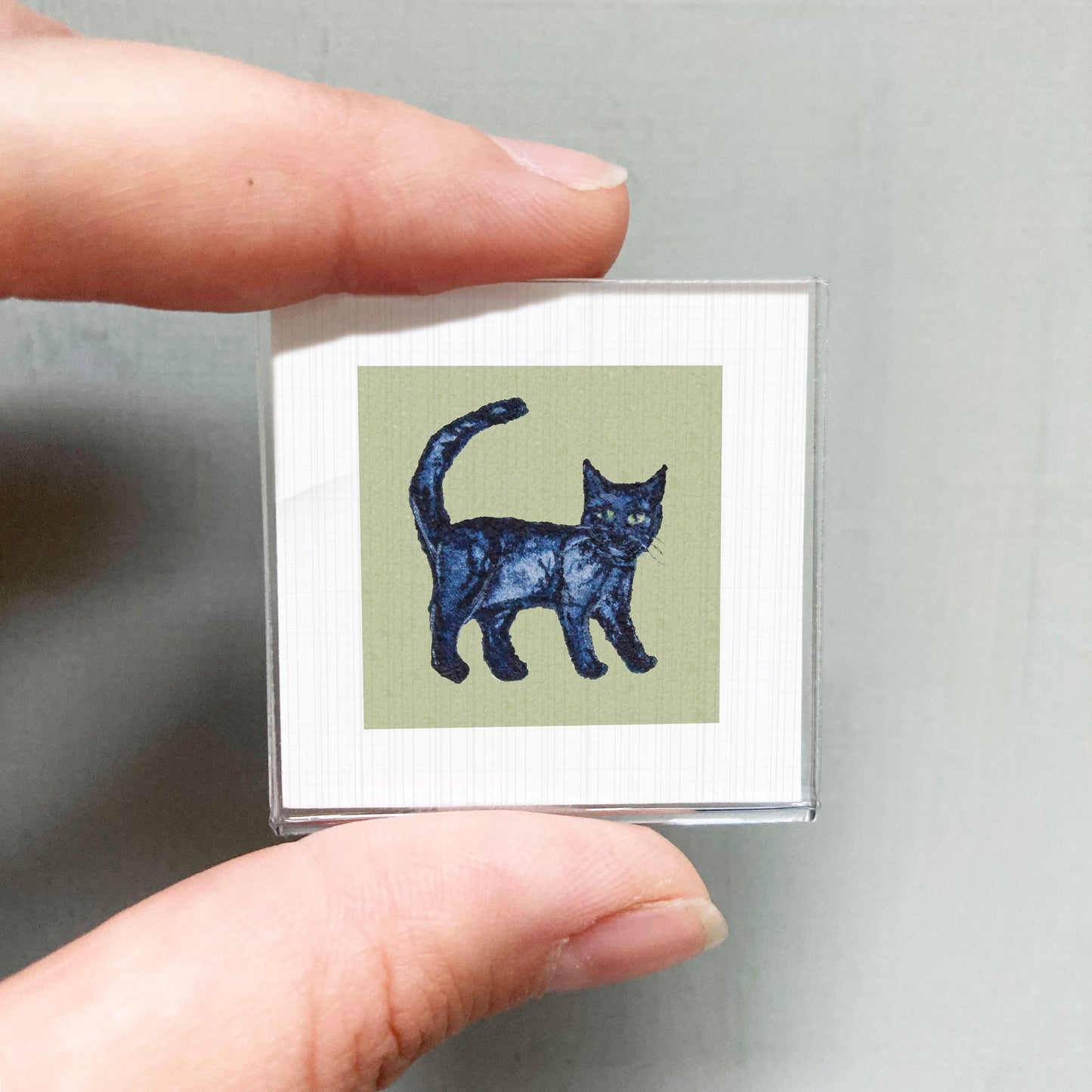 A person's hand is holding a small square with a black cat illustration on it.