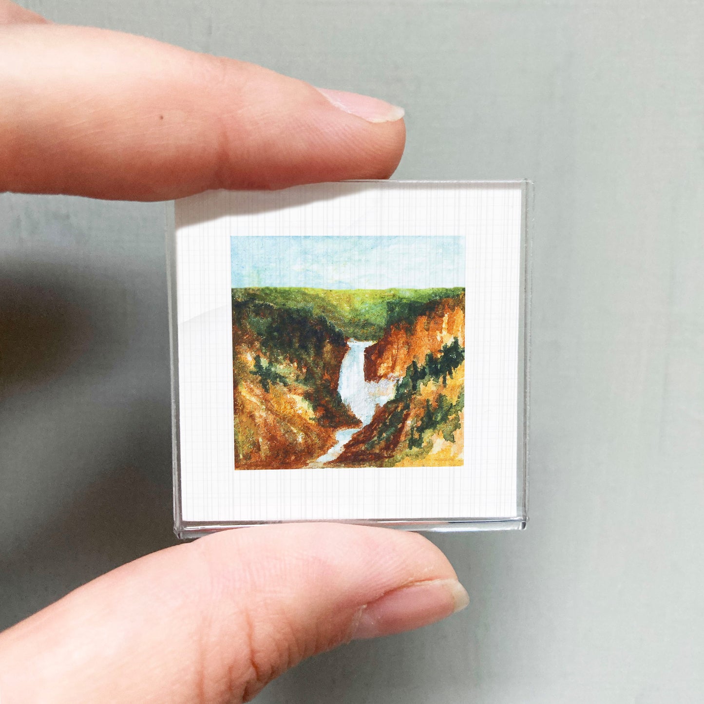 A person's hand holding a small square picture of a waterfall in a mountainous landscape.