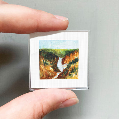 A person's hand holding a small square picture of a waterfall in a mountainous landscape.