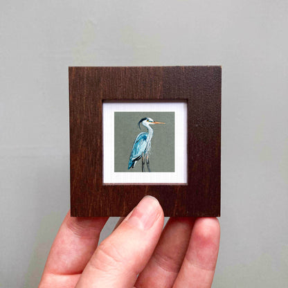 A person's hand holding a small wooden frame containing a painting of a blue heron.