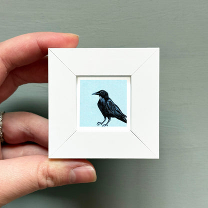 A hand is holding a small white square frame containing a black bird illustration.