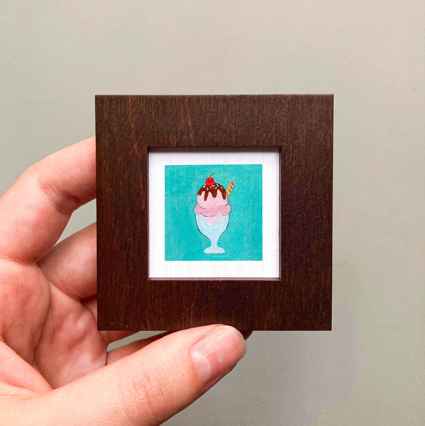 A hand is holding a small square picture frame containing an illustration of a colorful ice cream cone.
