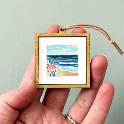 A hand holding a small wooden frame containing a painting of a beach scene with umbrellas and the ocean.