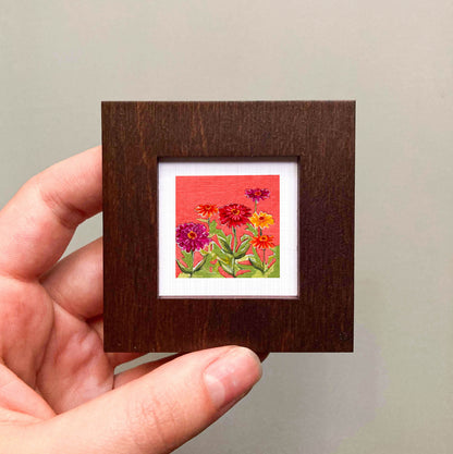 A hand is holding a small wooden frame containing a vibrant painting of colorful flowers.