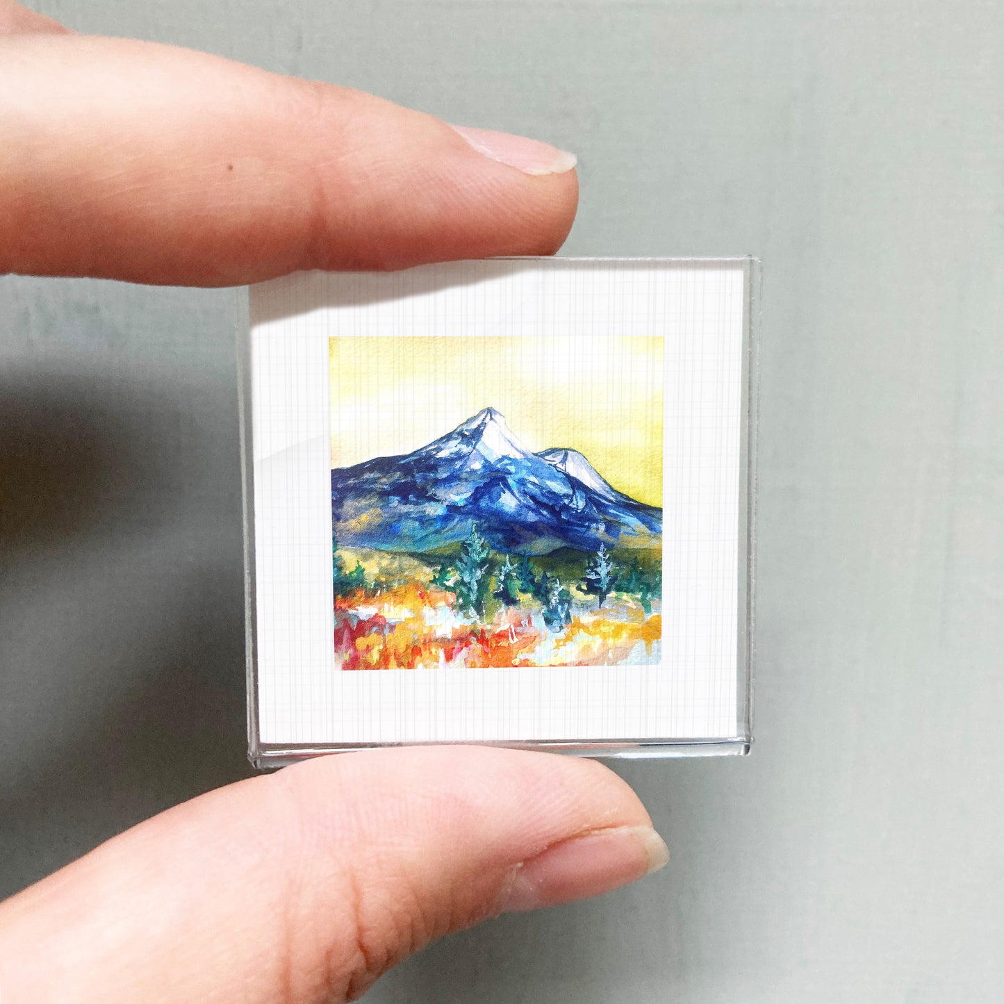 A person's hand holding a small square picture of a mountain landscape with a colorful, vibrant scene.