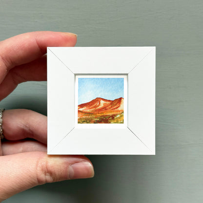 A hand is holding a small white square frame containing a watercolor painting of a mountain landscape.