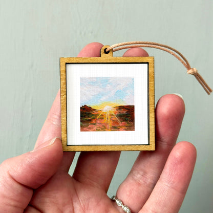 A hand holding a small wooden frame containing a painting of a landscape with a sunset.