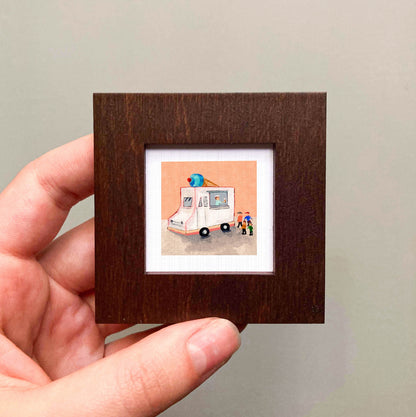 A hand is holding a small, square picture frame containing an illustration of an ice cream truck.