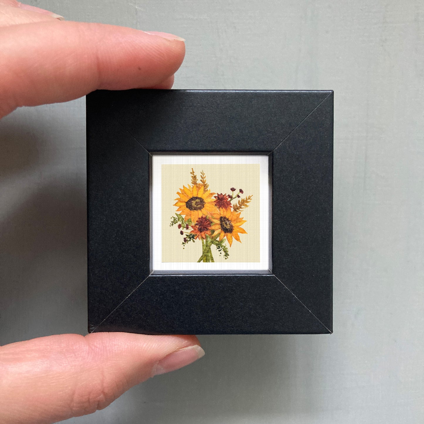 A person's hand holding a small black picture frame containing a framed image of a bouquet of sunflowers.