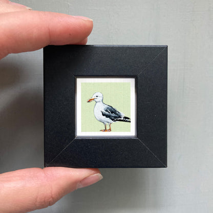 A person's hand holding a small black frame containing a framed image of a seagull.
