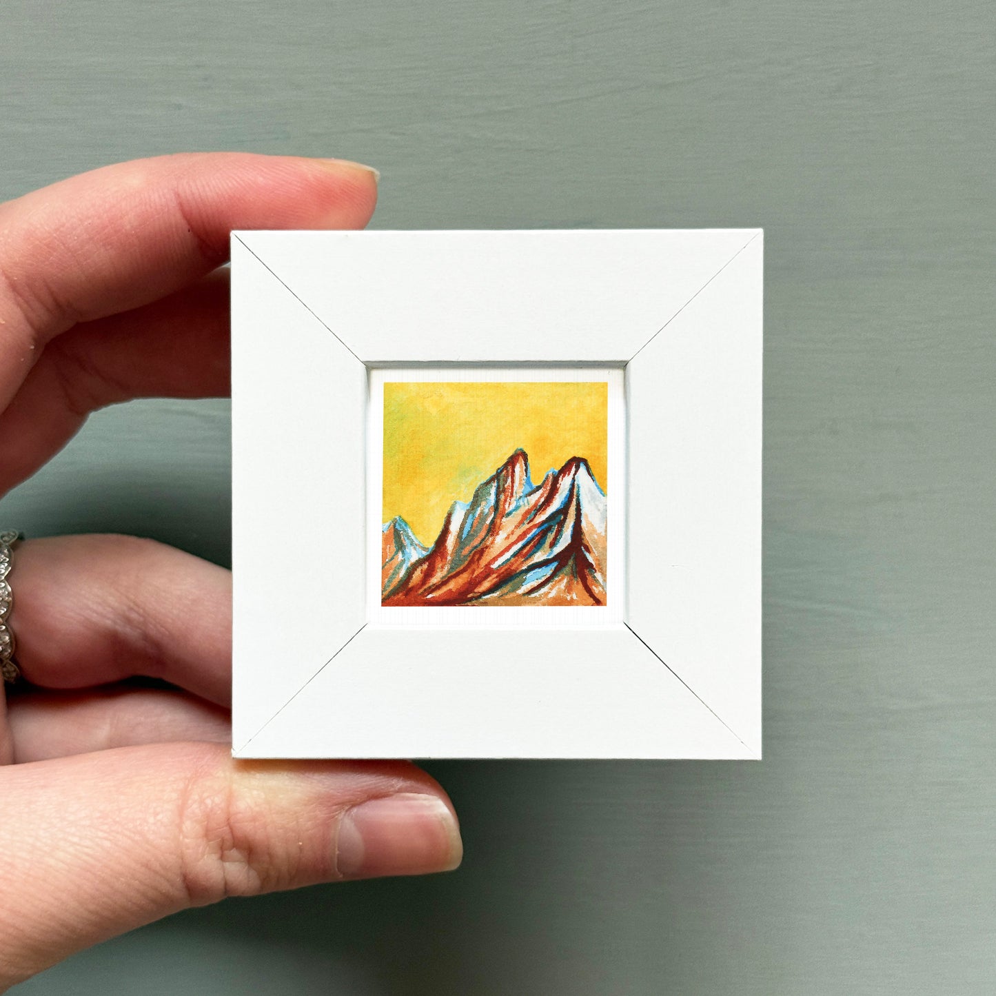 A hand is holding a small white square frame containing a colorful painting of mountains.