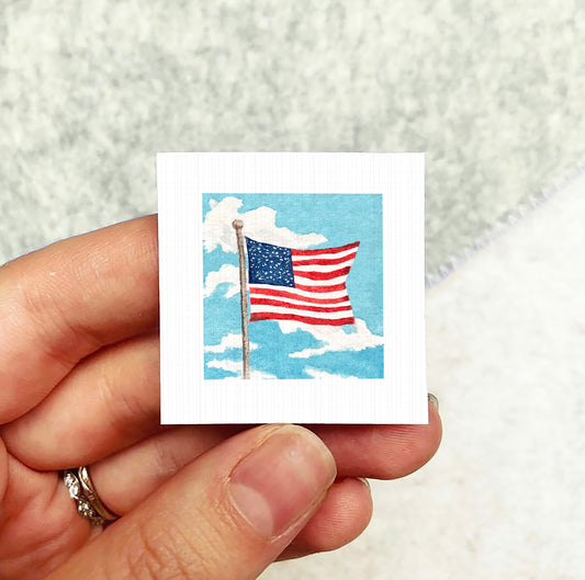 A hand holding a small square-shaped paper with an American flag on it against a cloudy sky background.
