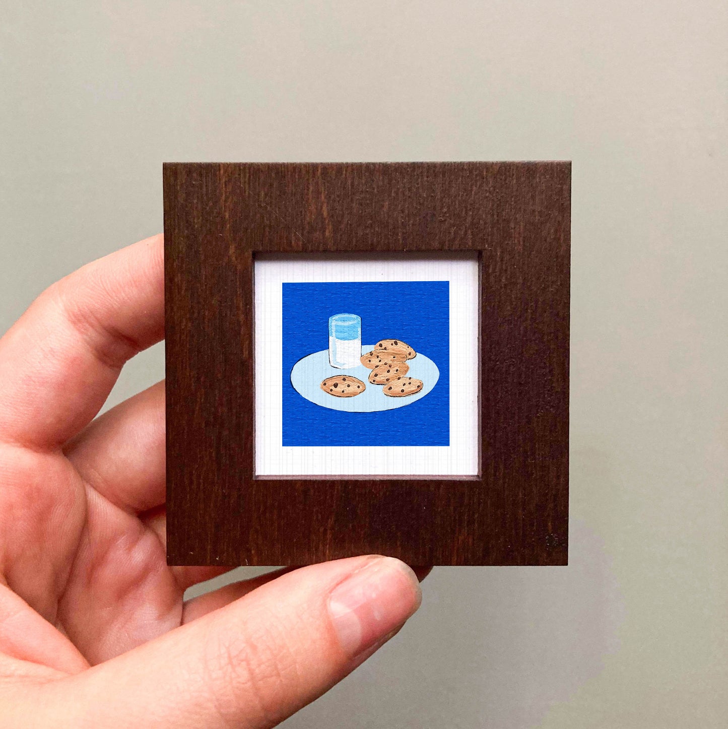 A hand is holding a small wooden frame containing a framed illustration of cookies and a glass of milk.