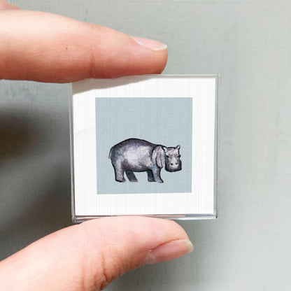 A person's hand is holding a small square picture of a hippopotamus against a light blue background.