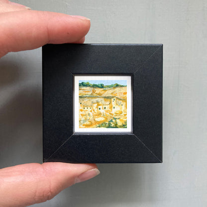 A person's hand holding a small black frame containing a miniature painting of a landscape.
