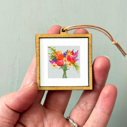 A hand holding a small wooden frame containing a framed painting of a bouquet of colorful flowers.